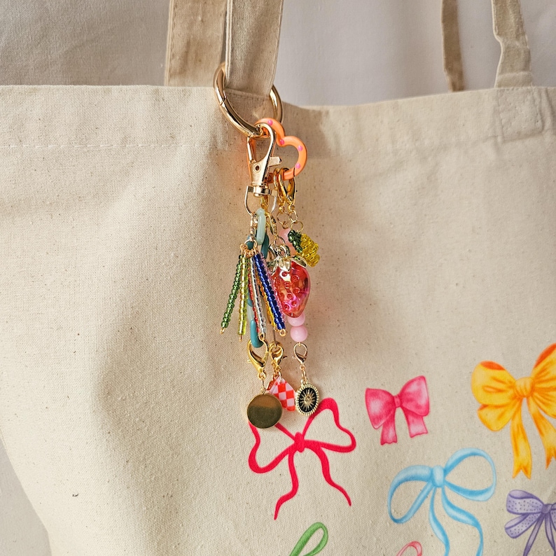 May include: A gold key chain with a heart charm, beads, and a strawberry charm. The key chain is attached to a white tote bag with a pink, yellow, and blue bow design.