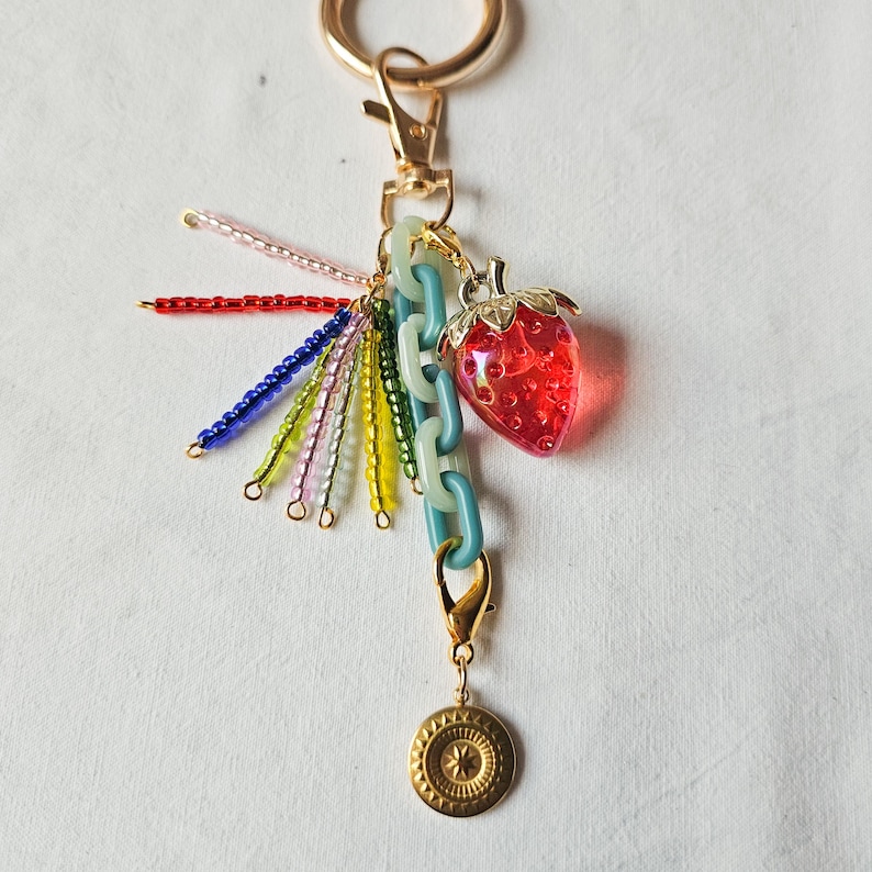 May include: A gold key chain with a red and clear plastic strawberry charm, a blue, green, red, and white beaded tassel, and a gold circular charm with a star design.