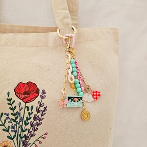 May include: A beige canvas tote bag featuring a floral print of a red poppy and purple lavender. Attached to the handle is a multi-strand charm keychain with a pink butterfly, a teal cassette tape, beads, a white cloud, and a red checkered heart.