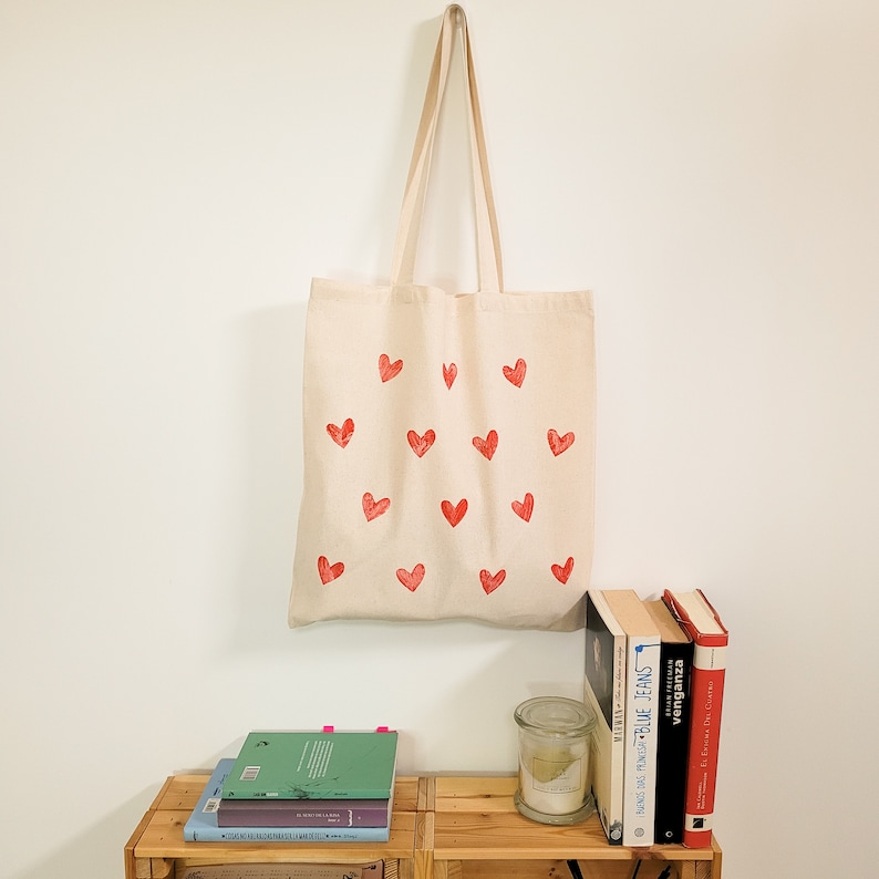 May include: A beige tote bag with red heart designs printed on the front. The bag is hanging on a white wall with a stack of books and a glass jar on a wooden shelf below.