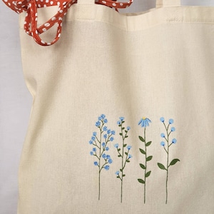 Peut inclure: Un sac fourre-tout en toile blanche avec un motif peint &agrave; la main de fleurs bleues et de tiges vertes. Les fleurs sont dispos&eacute;es en ligne verticale au centre du sac.