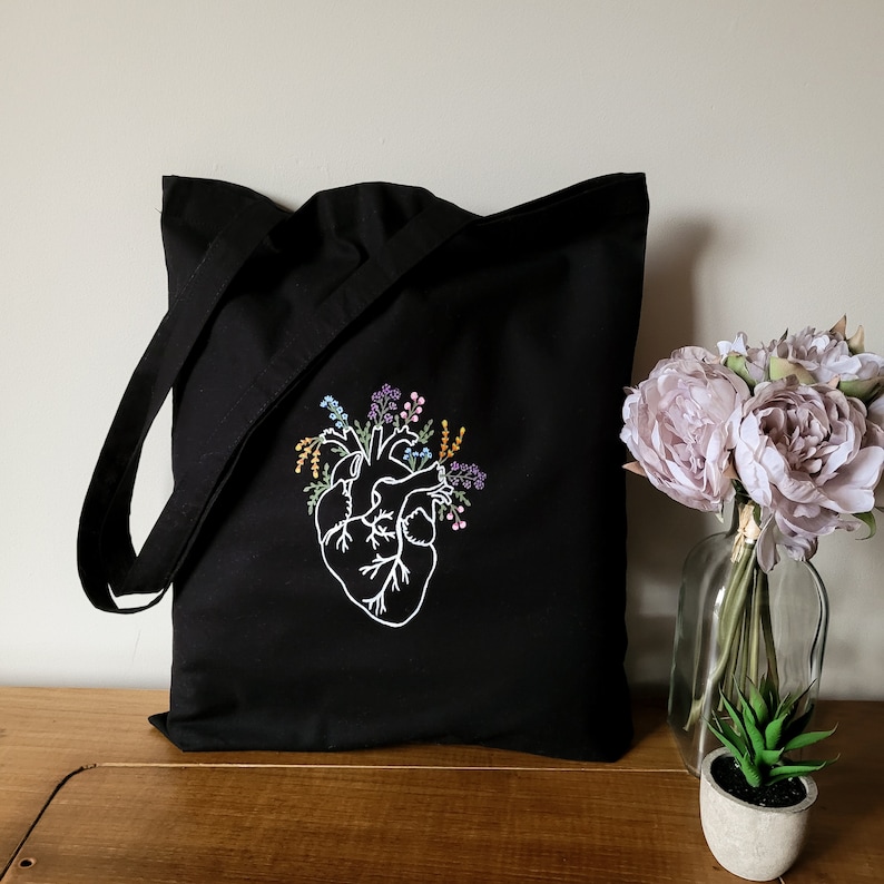 K&ouml;nnte beinhalten: Schwarze Tragetasche mit einem wei&szlig;en gestickten anatomischen Herzen mit floralen Details. Das Herz ist von kleinen Blumen in den Farben Lila, Gelb und Orange umgeben.