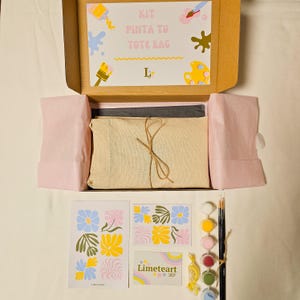 May include: A tote bag painting kit in an open cardboard box. The kit includes a canvas tote bag, paints, brushes, and a design guide. The box lid has the text "KIT PINTA TU TOTE BAG". Additional items include a Limeteart Shop card and floral design cards.