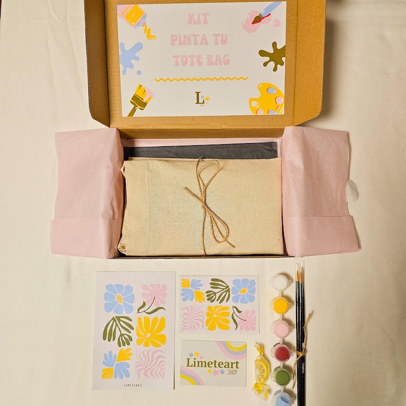 May include: A tote bag painting kit in an open cardboard box. The kit includes a canvas tote bag, paints, brushes, and a design guide. The box lid has the text "KIT PINTA TU TOTE BAG". Additional items include a Limeteart Shop card and floral design cards.