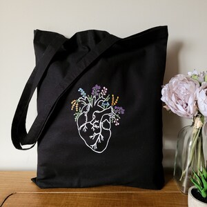 K&ouml;nnte beinhalten: Schwarze Tragetasche mit einem wei&szlig;en gestickten anatomischen Herzen, aus dem bunte Blumen wachsen.