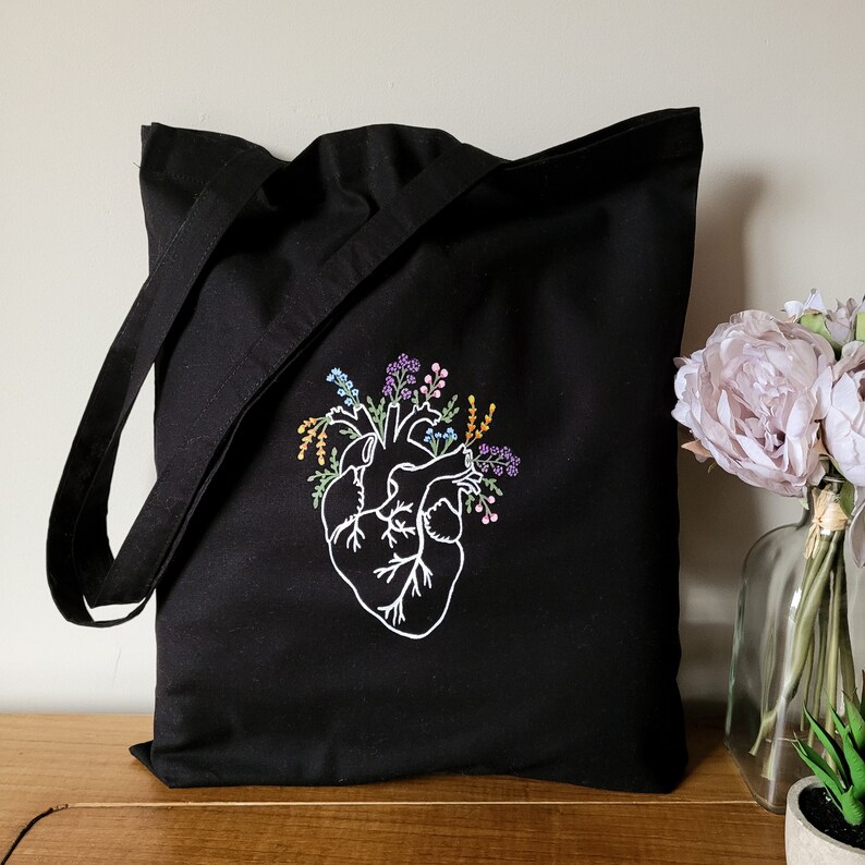 K&ouml;nnte beinhalten: Schwarze Tragetasche mit einem wei&szlig;en gestickten anatomischen Herzen, aus dem bunte Blumen wachsen.