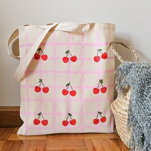 May include: A white canvas tote bag with pink and white checkered pattern. The bag is decorated with red cherries printed on the front.