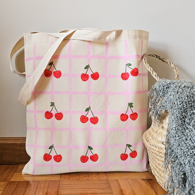 May include: A white canvas tote bag with pink and white checkered pattern. The bag is decorated with red cherries printed on the front.