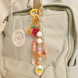 Peut inclure: Un porte-clés doré avec divers breloques, dont une fraise rouge, un soleil, un champignon et des perles colorées. Le porte-clés est attaché à un sac à dos vert clair. Les breloques sont de formes et de couleurs variées.