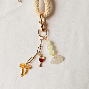 Puede incluir: Un llavero dorado con un dije de copa de vino, un dije de nube y una cadena de cuentas verdes. El llavero est&aacute; unido a una cuerda blanca.