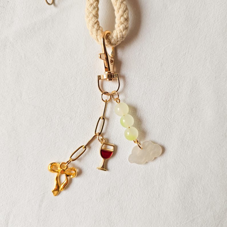 Puede incluir: Un llavero dorado con un dije de copa de vino, un dije de nube y una cadena de cuentas verdes. El llavero est&aacute; unido a una cuerda blanca.