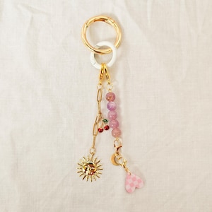 May include: A gold-colored keychain with a large ring and a white accent ring. The keychain features a chain, a sun charm, a cherry charm, a strand of purple beads, a crescent moon charm, and a pink checkered heart charm.