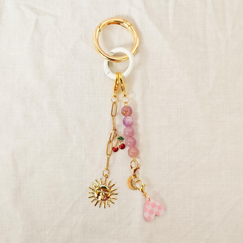 May include: A gold-colored keychain with a large ring and a white accent ring. The keychain features a chain, a sun charm, a cherry charm, a strand of purple beads, a crescent moon charm, and a pink checkered heart charm.
