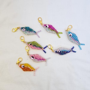 May include: A collection of beaded fish charms in various colors, including blue, pink, green, and yellow. Each charm has a gold-colored clasp. The fish are detailed with beadwork, featuring fins and eyes.