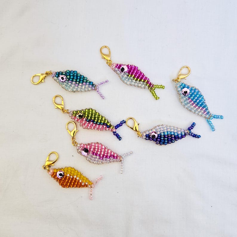 May include: A collection of beaded fish charms in various colors, including blue, pink, green, and yellow. Each charm has a gold-colored clasp. The fish are detailed with beadwork, featuring fins and eyes.