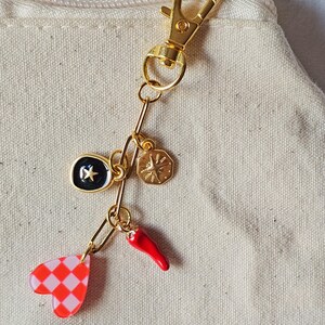 May include: Gold-toned keychain with multiple charms. Charms include a black and gold star, a gold compass, a red chili pepper, and a pink and red checkered heart. The keychain is attached to a beige fabric.