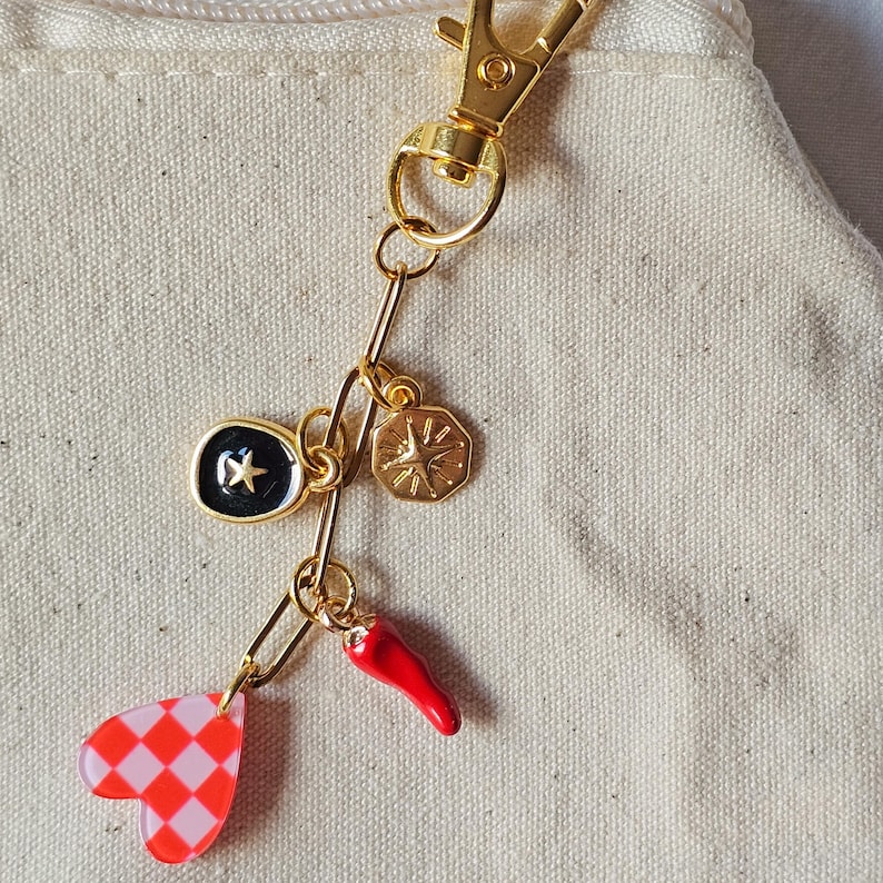 May include: Gold-toned keychain with multiple charms. Charms include a black and gold star, a gold compass, a red chili pepper, and a pink and red checkered heart. The keychain is attached to a beige fabric.