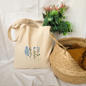Peut inclure: Un sac fourre-tout en toile blanche avec une broderie florale bleue. Le sac a deux anses et est pos&eacute; sur une surface blanche. Un panier en osier est en arri&egrave;re-plan.