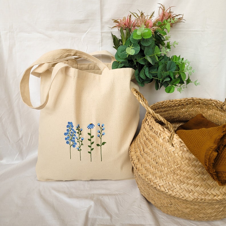 Peut inclure: Un sac fourre-tout en toile blanche avec une broderie florale bleue. Le sac a deux anses et est pos&eacute; sur une surface blanche. Un panier en osier est en arri&egrave;re-plan.
