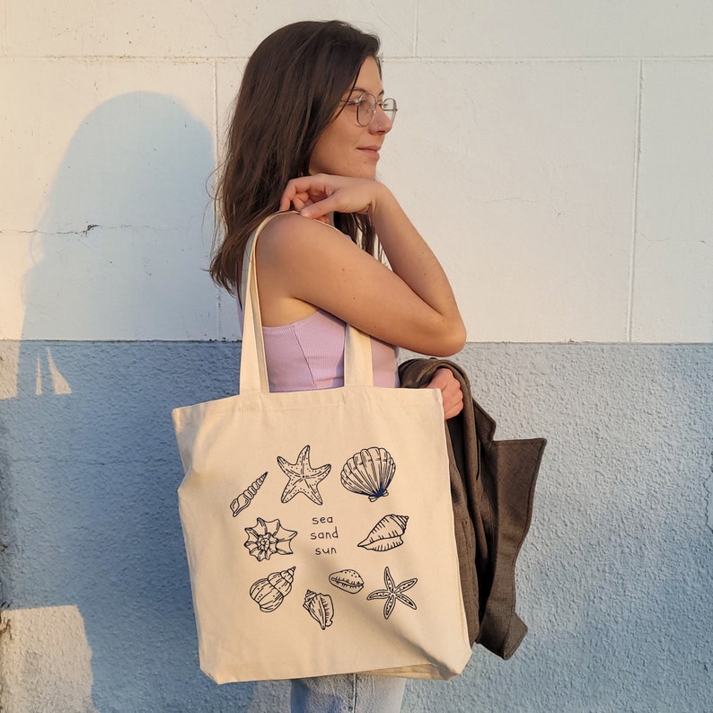 May include: A beige tote bag with a black and white illustration of seashells and starfish. The bag has the text "sea sand sun" printed on it.