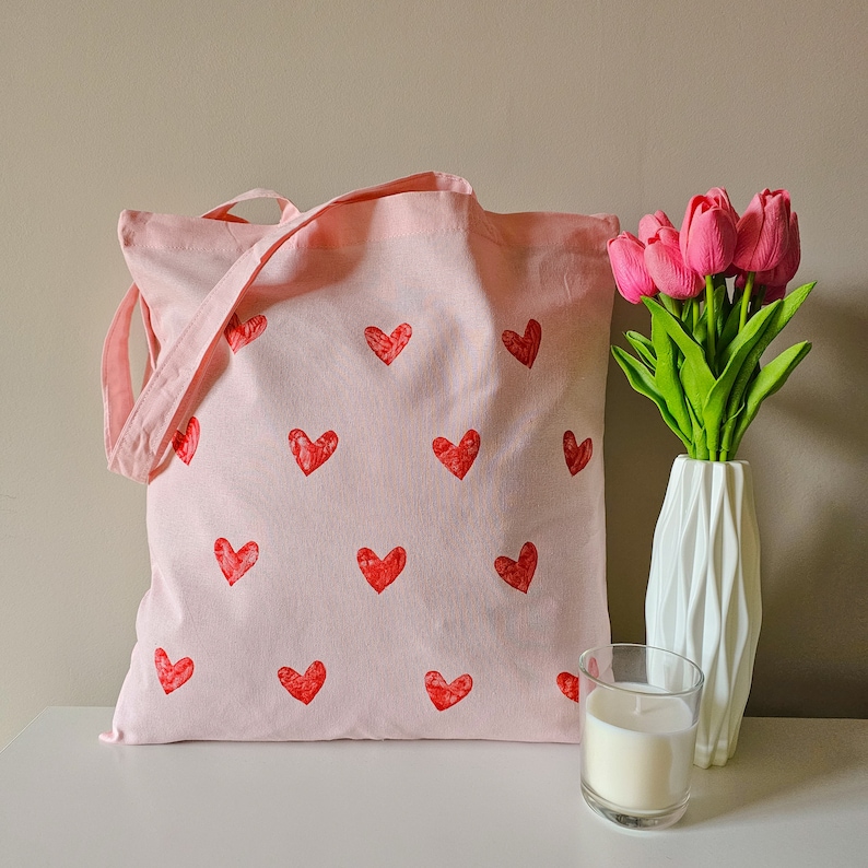 May include: A pink canvas tote bag with red heart designs. The bag has a long strap and is made of a soft, durable fabric.