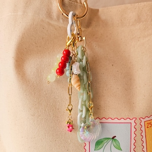 以下が含まれることがあります： 赤いビーズと緑のビーズ、貝殻、ピンクの花など、さまざまなチャームが付いたゴールドのキーチェーン。チャームはゴールドチェーンと白いプラスチックチェーンに取り付けられています。