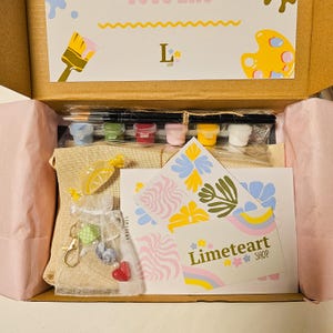 May include: An open box reveals a paint-by-numbers kit with paint pots, brushes, and a canvas. The kit includes a small bag of candies and a keychain. The box also contains cards with the text "Limeteart SHOP" and decorative floral designs.