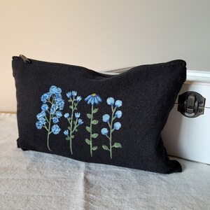 May include: Black canvas zippered pouch with embroidered blue flowers. The flowers are in a row and include a daisy and forget-me-nots.
