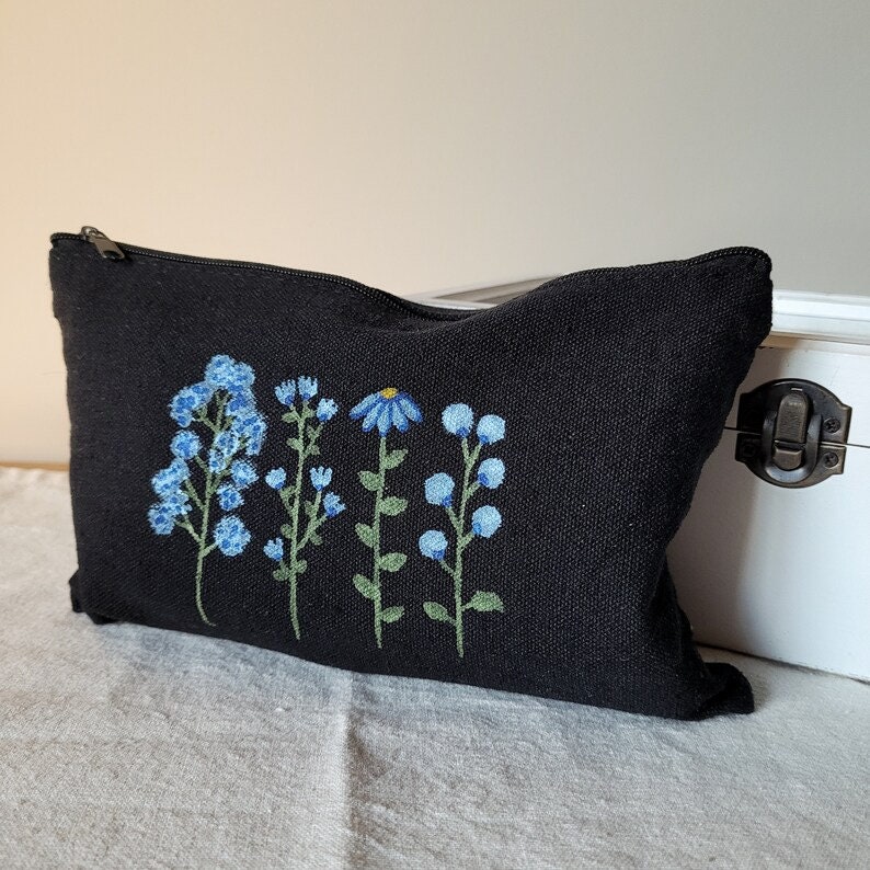 May include: Black canvas zippered pouch with embroidered blue flowers. The flowers are in a row and include a daisy and forget-me-nots.