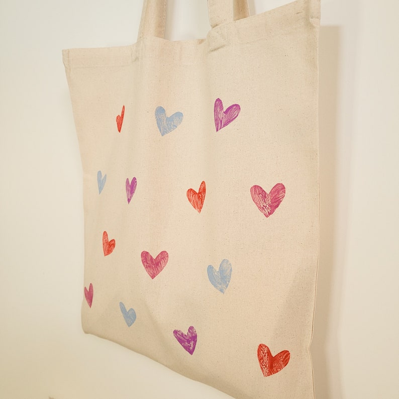 May include: A beige canvas tote bag with a variety of colorful heart designs printed on it. The hearts are in shades of pink, red, purple, and blue.