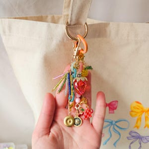 May include: A colorful charm with a strawberry, beads, and a gold chain attached to a gold key ring. The charm is hanging from a white canvas tote bag.