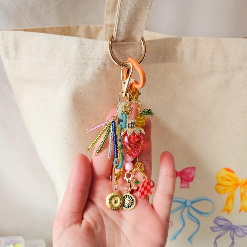 May include: A colorful charm with a strawberry, beads, and a gold chain attached to a gold key ring. The charm is hanging from a white canvas tote bag.