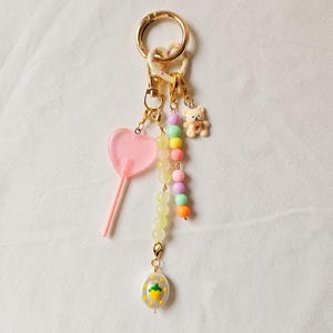 Può includere: Un portachiavi dorato con un ciondolo a forma di lecca-lecca a cuore rosa, perline color pastello, un piccolo ciondolo a forma di orsacchiotto e un ciondolo a fetta di limone.