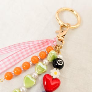 May include: A beaded keychain with a gold-colored clasp and ring. The keychain features orange beads, green heart-shaped beads, white pearls, and a black 8-ball charm. A red heart and a white daisy charm are also included.