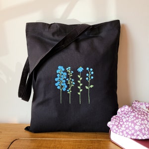 K&ouml;nnte beinhalten: Schwarze Tragetasche mit einem Design aus vier blauen Blumen, die auf der Vorderseite gestickt sind. Die Blumen sind stilisiert und haben gr&uuml;ne Stiele.