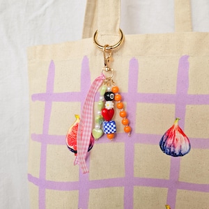 May include: A canvas tote bag with a gold-colored keyring and a beaded charm. The charm includes an 8-ball bead, heart beads, and orange beads. The bag has a purple grid pattern and fig illustrations.