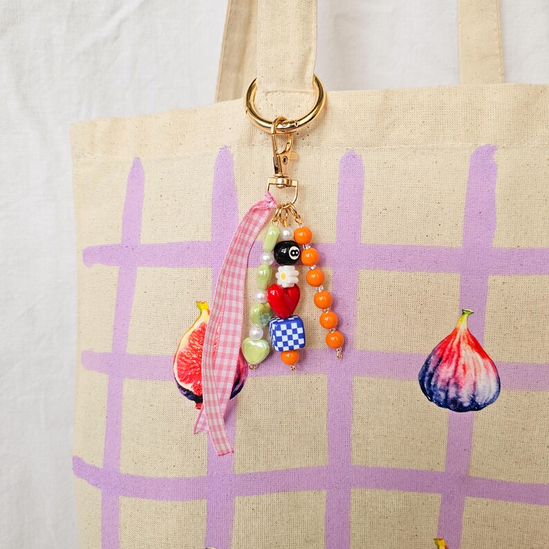 May include: A canvas tote bag with a gold-colored keyring and a beaded charm. The charm includes an 8-ball bead, heart beads, and orange beads. The bag has a purple grid pattern and fig illustrations.