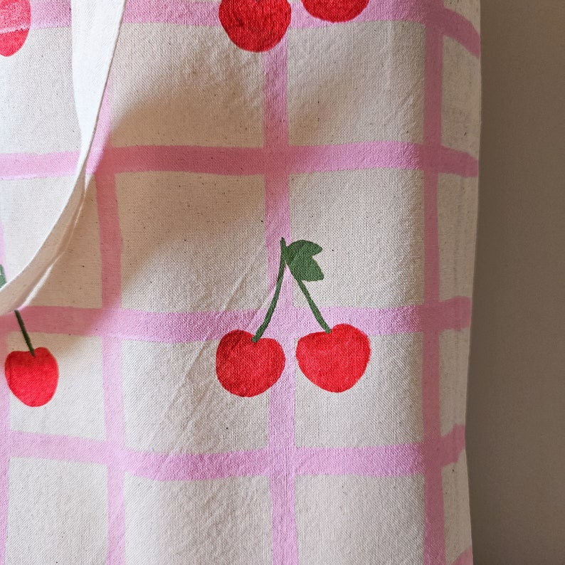 May include: A white canvas tote bag with pink checkered pattern and red cherries painted on it.