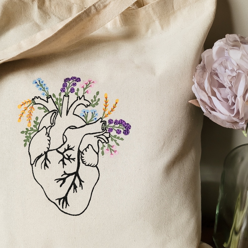 K&ouml;nnte beinhalten: Eine wei&szlig;e Canvas-Tragetasche mit einem gestickten Design eines menschlichen Herzens, aus dem Blumen wachsen. Die Blumen sind in den Farben Lila, Rosa, Gelb und Blau.