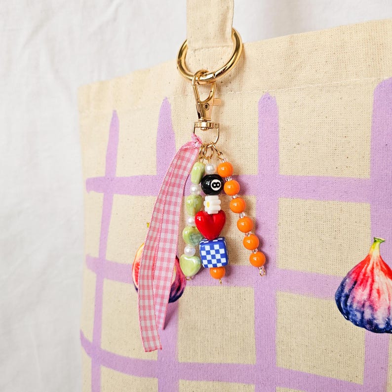 May include: A gold-toned keychain with a beaded charm, attached to a canvas tote bag. The charm features a variety of beads, including a black 8-ball, orange spheres, and a red heart. A pink gingham ribbon is also attached.