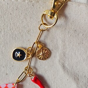 May include: A gold-colored keychain with a lobster clasp and decorative charms. Charms include a black oval with a gold star, a gold sunburst, and a red chili pepper. The chain is made of gold-colored links.