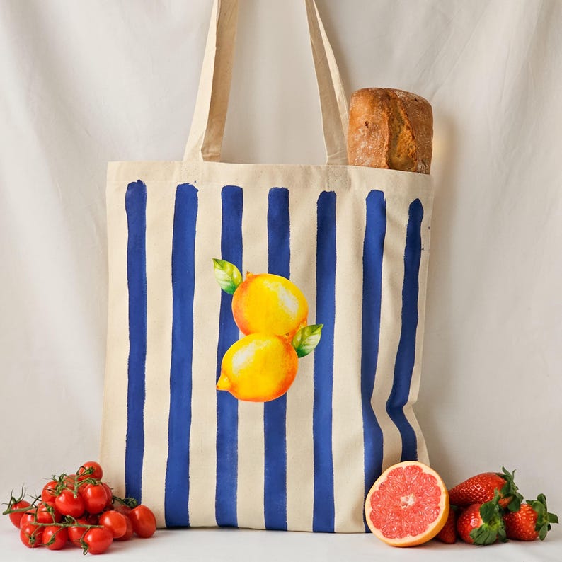 May include: A white canvas tote bag with blue stripes and a lemon design. The bag is filled with a loaf of bread and has a red grapefruit and strawberries next to it.