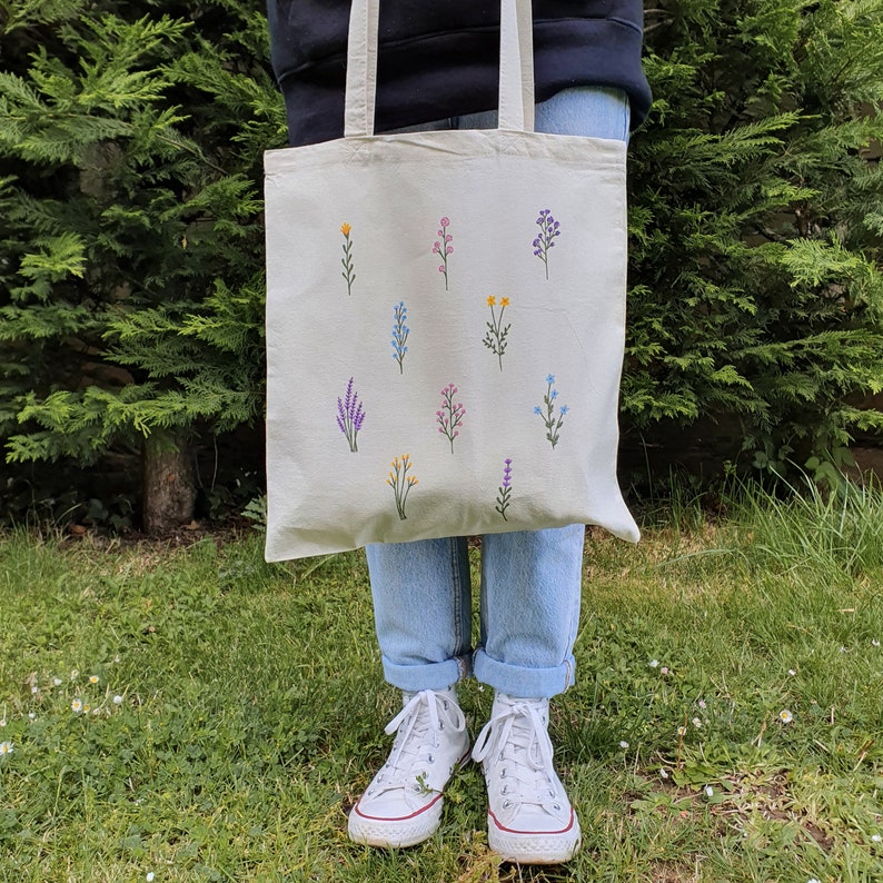 May include: A white canvas tote bag with embroidered flowers in various colors, including purple, pink, blue, and yellow. The bag has two long straps and is being held by a person wearing blue jeans and white sneakers.