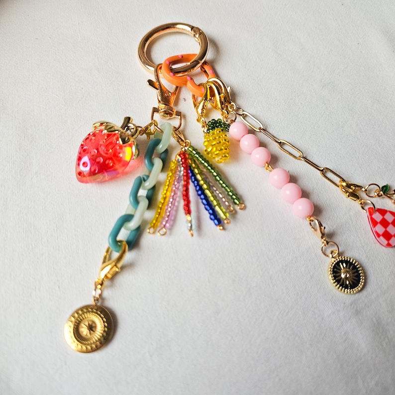May include: A gold key chain with a variety of charms, including a red strawberry, a pineapple, a pink bead, a green and white checkered charm, and a gold sun charm.