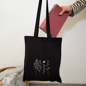 May include: Black tote bag with a floral design embroidered on the front. The flowers are purple, blue, white, and red. The bag is being held by a hand holding a red book.
