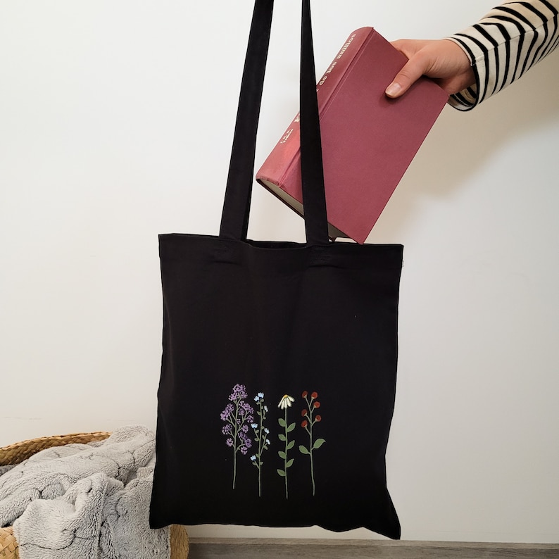 May include: Black tote bag with a floral design embroidered on the front. The flowers are purple, blue, white, and red. The bag is being held by a hand holding a red book.