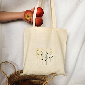 Puede incluir: Una bolsa de lona blanca con un diseño floral que presenta cuatro tallos verdes con flores amarillas. La bolsa tiene dos asas y se muestra con tres melocotones rojos.