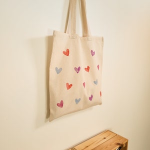 May include: A beige tote bag with a pattern of red, pink, and blue hearts. The bag has two straps and is made of canvas.
