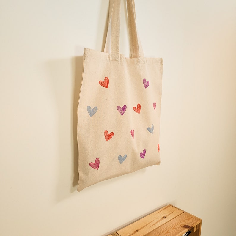 May include: A beige tote bag with a pattern of red, pink, and blue hearts. The bag has two straps and is made of canvas.