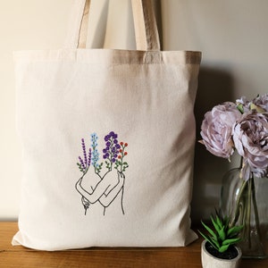 May include: A white canvas tote bag with a black line drawing of two figures embracing. The figures have flowers growing from their heads, including purple, blue, red, and green flowers.